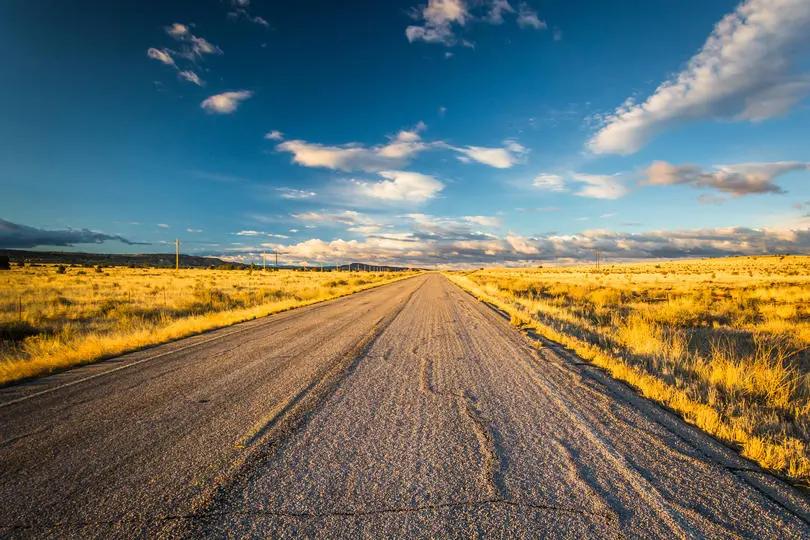 View of a straight rural road cutting through golden fields under a dramatic sky with scattered clouds at sunset.
