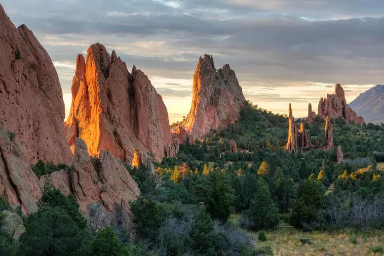 View of dramatic red rock formations glowing at sunrise, surrounded by lush greenery in a rugged canyon landscape.