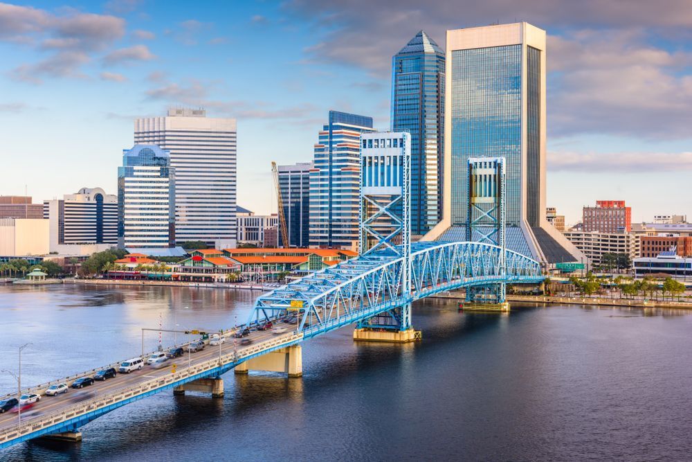 Vue de la silhouette de Jacksonville avec le pont Green Monster bleu franchissant la St. Johns River, entourée d’immeubles modernes et de paysages urbains sous un ciel clair.