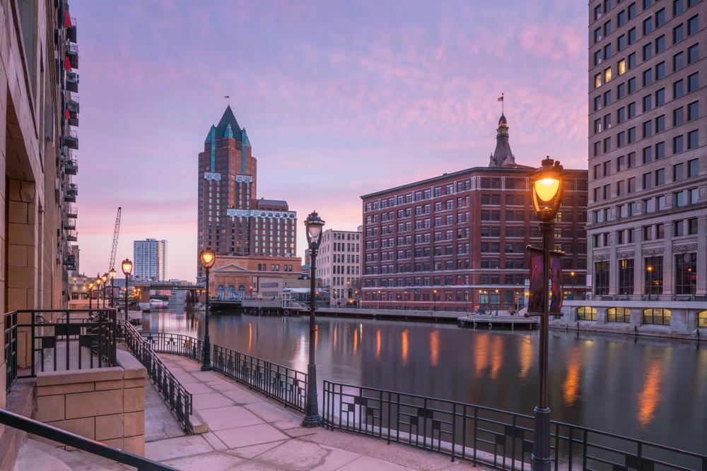 Downtown Milwaukee Riverwalk at sunset with historic buildings, street lamps, and calm water reflections.