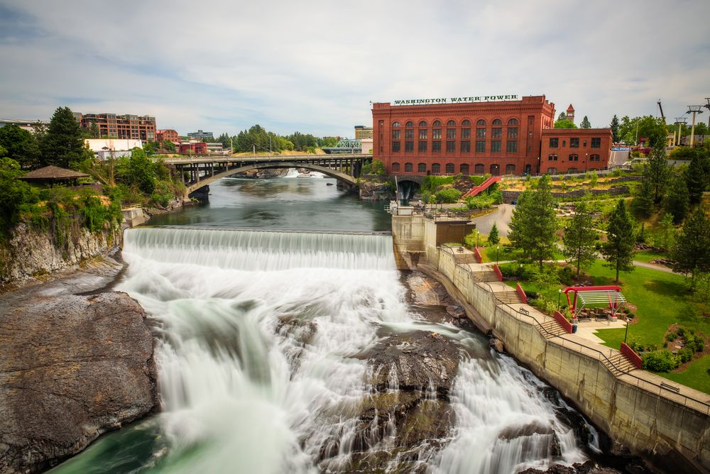 Urban waterfall flowing through a city park, with a historic red brick power building and a bridge in the background.