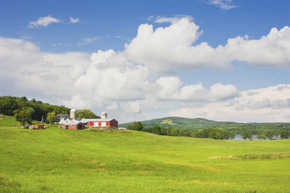 Scenic rural landscape with green pastures, a red barn, silos, and rolling hills beneath a partly cloudy sky.