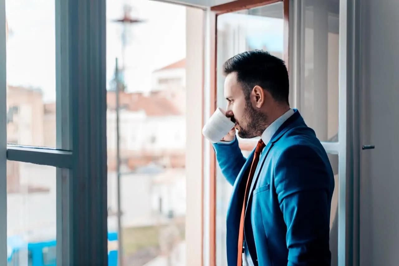 View of a man in a blue suit drinking coffee while looking out of a window in a modern urban setting.