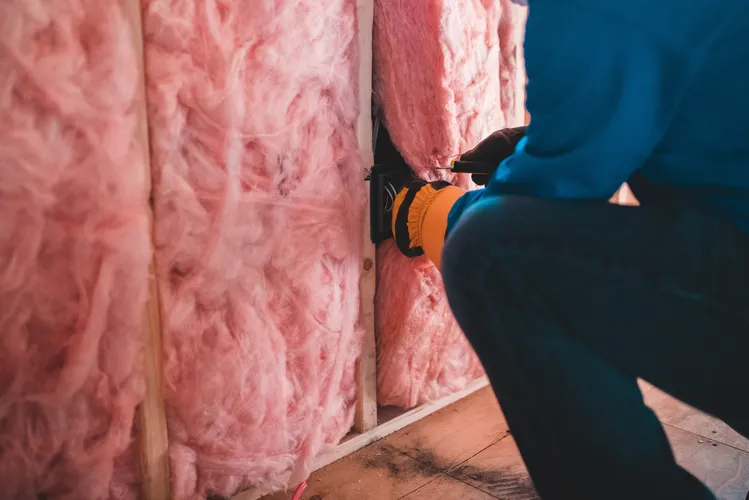 A view of a worker installing electrical outlet in wall with pink fiberglass insulation.