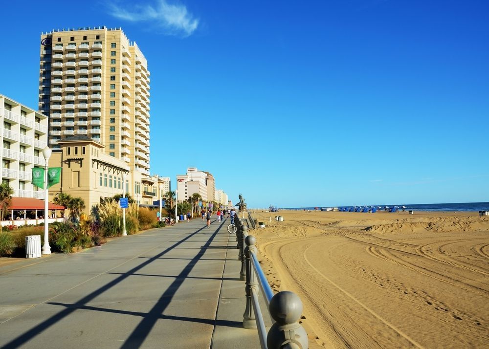 Vue d’une promenade en bord de mer avec des immeubles en arrière‑plan, des personnes marchant et faisant du vélo, et une plage bordée de parasols sous un ciel bleu clair.