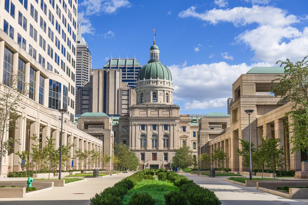 Indiana State Capitol building in downtown Indianapolis with blue skies and surrounding modern architecture.