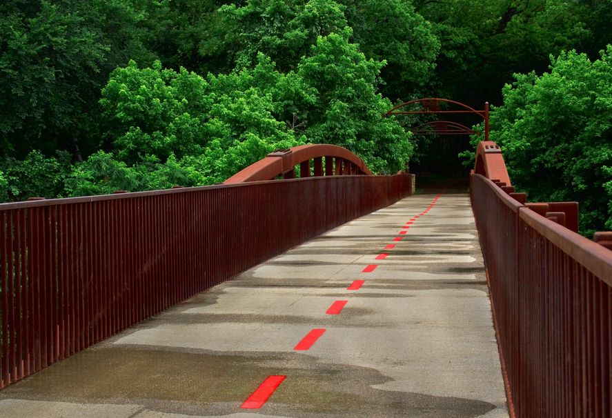 A view of a red-painted pedestrian bridge, with green trees surrounding the path and a serene, natural environment in the background. The bridge is wet, possibly after rain, with a line of red marking the walkway.