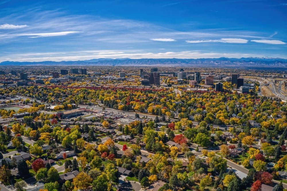Vue aérienne d’une ville entourée d’arbres d’automne colorés, avec des teintes vives de rouge, d’orange et de jaune se mêlant au paysage urbain et aux montagnes au loin sous un ciel bleu clair.