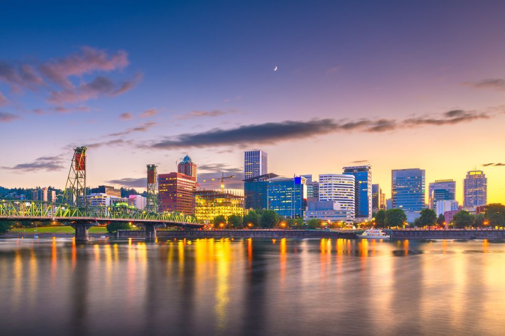 Vue pittoresque de la silhouette de Portland au crépuscule, avec la Willamette reflétant les lumières de la ville et le pont vert emblématique au premier plan. La ville est baignée de tons chauds du coucher de soleil.