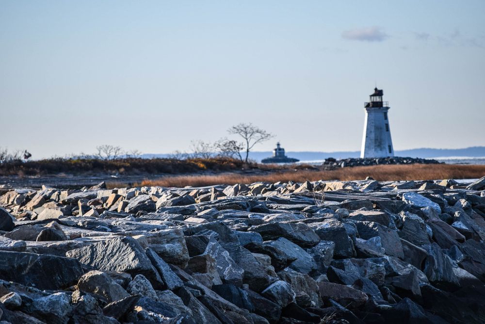 Stone jetty leading to a lighthouse with a calm coastal backdrop under clear skies.