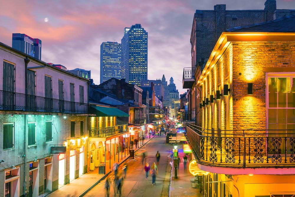 A view of Bourbon Street in New Orleans at dusk with colorful lights, historic French Quarter architecture, and the city skyline in the background.