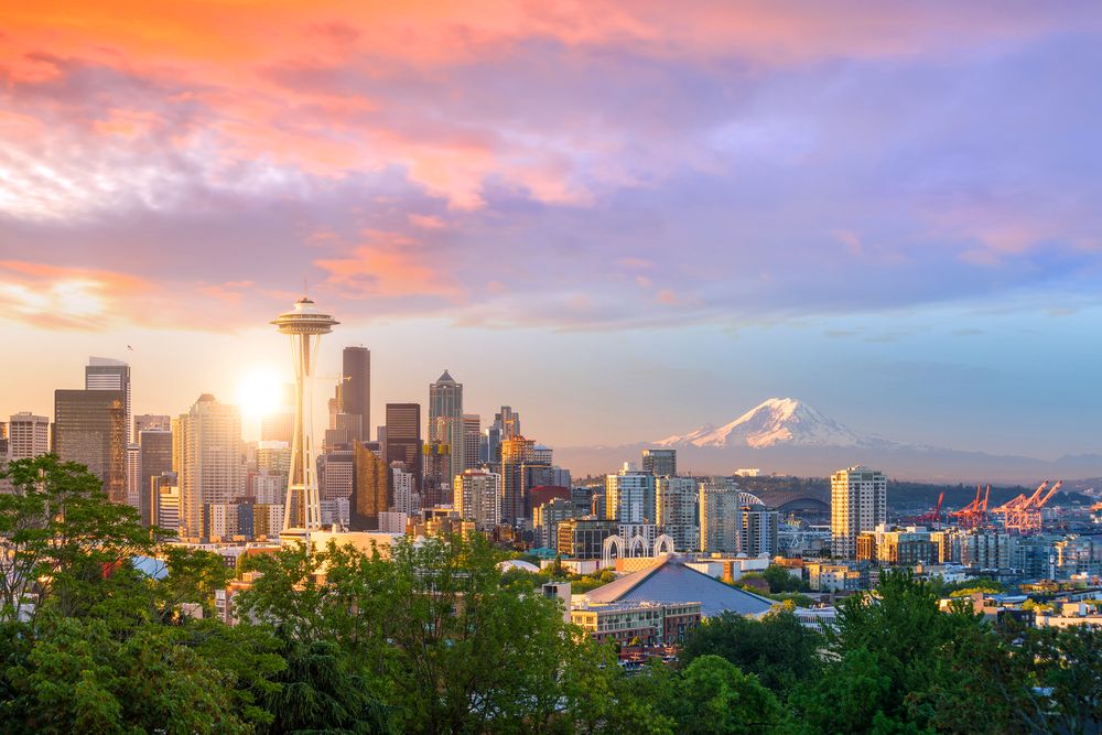 A scenic view of Seattle's skyline at sunset, featuring the Space Needle and Mount Rainier in the distance, with vibrant colors filling the sky and illuminating the city.