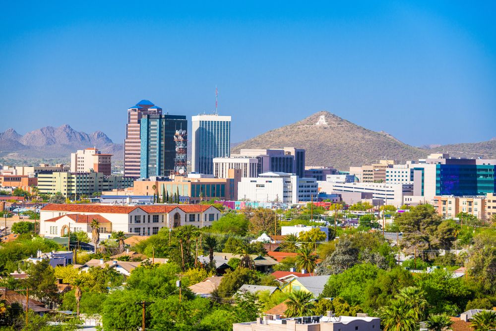 A view of Tucson, Arizona, with its modern cityscape featuring tall buildings and green spaces, set against a backdrop of desert mountains under a clear blue sky.