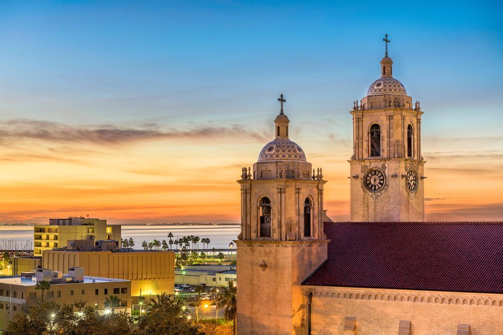 Vue de deux clochers d’église au coucher du soleil, avec une douce lueur orange et bleue dans le ciel, surplombant une ville côtière avec des palmiers et des bâtiments en bord de mer au loin.
