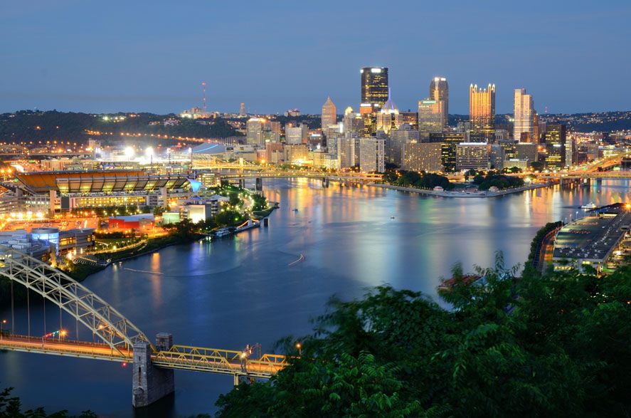 Pittsburgh’s illuminated skyline at night, seen from a distance, with bridges over the river, suggesting a city where industrial history may impact air quality