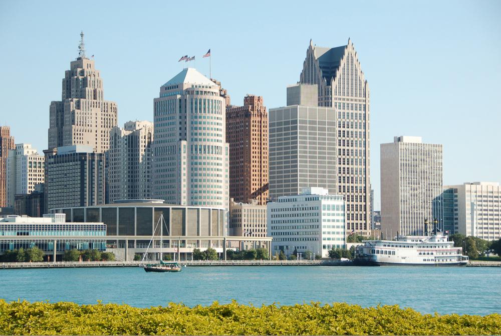 Detroit skyline featuring iconic skyscrapers along the waterfront under a clear sky, viewed from across the river.