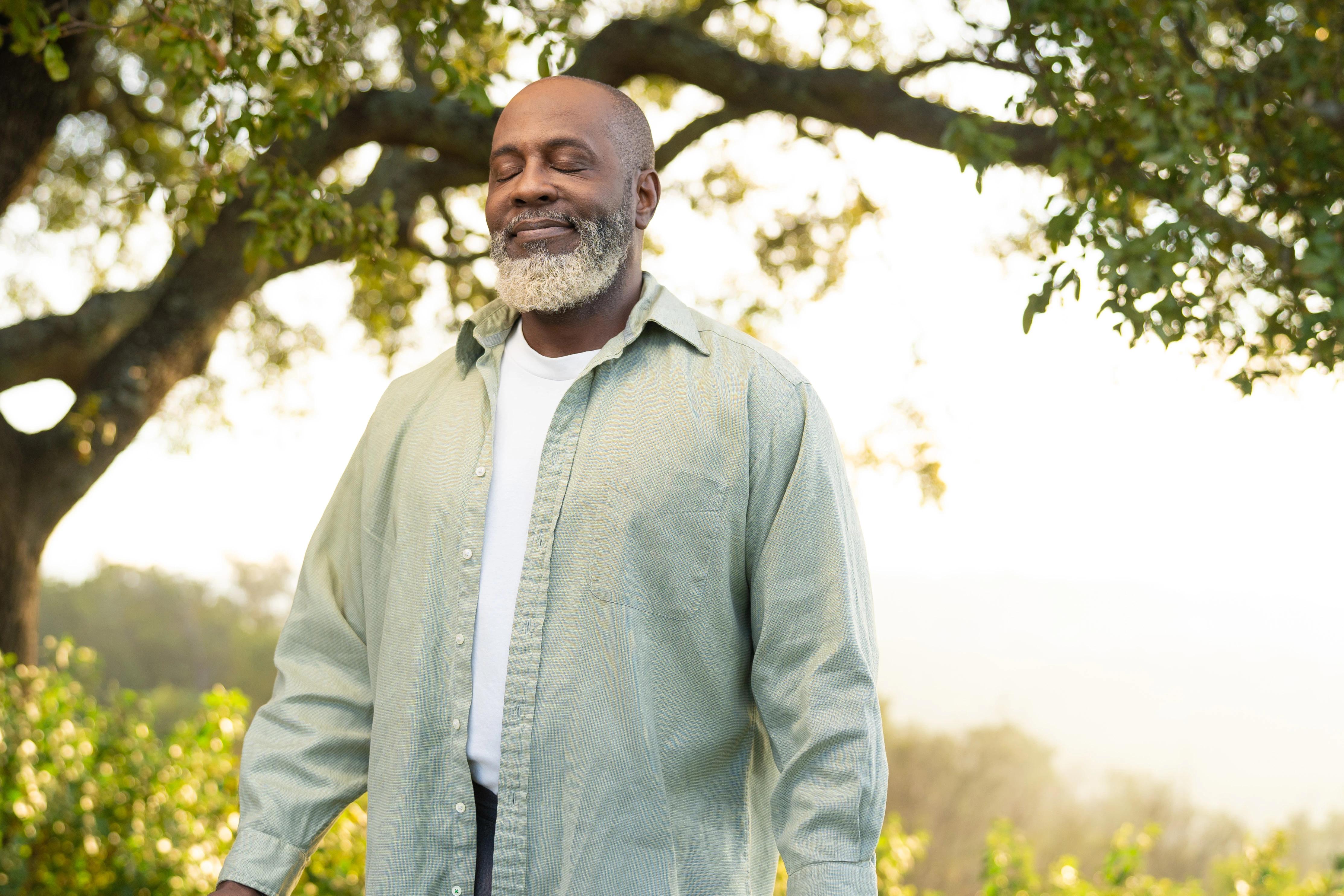 A mature man standing peacefully outdoors with eyes closed, enjoying fresh air and sunlight under a large oak tree.