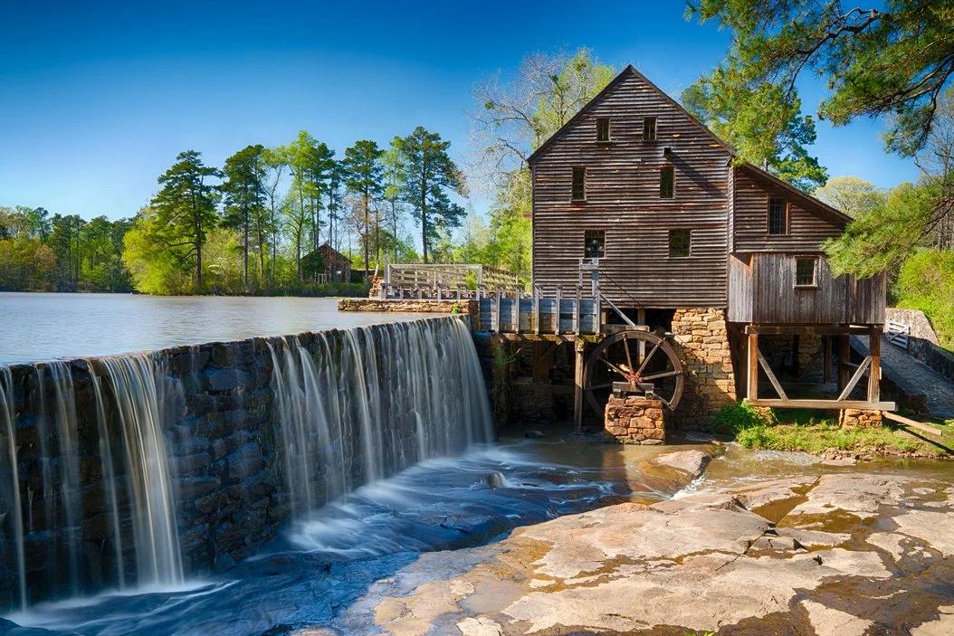 View of a historic wooden watermill beside a stone dam with water flowing over, surrounded by trees and a clear blue sky.