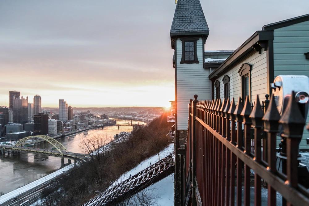 A view of Pittsburgh from the Duquesne Incline at sunrise, overlooking the city skyline, river bridges, and snow-covered hillside.