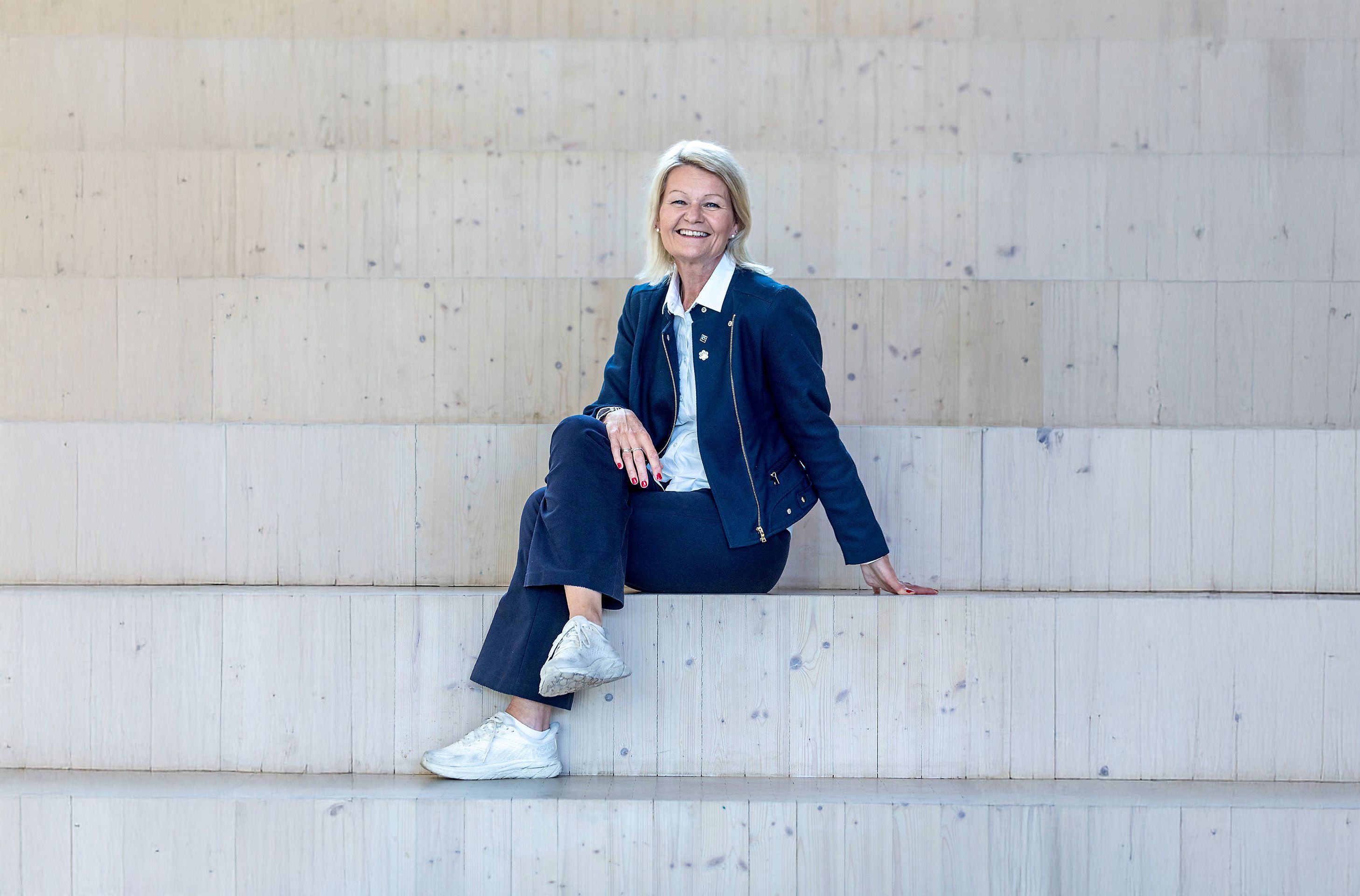 Rektor Anne Kristine Eggen Lervik fra Fagskolen Oslo sitter smilende på en tretrapp. Hun er kledd i en mørkeblå jakke og bukse, og hvite joggesko, mot en bakgrunn av lyse trepaneler.