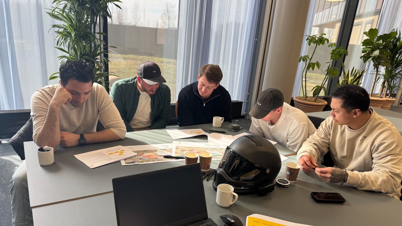 Fem studenter sitter rundt et bord i et opplyst rom og samarbeider om et prosjekt, med papirer og en laptop spredt utover bordet.