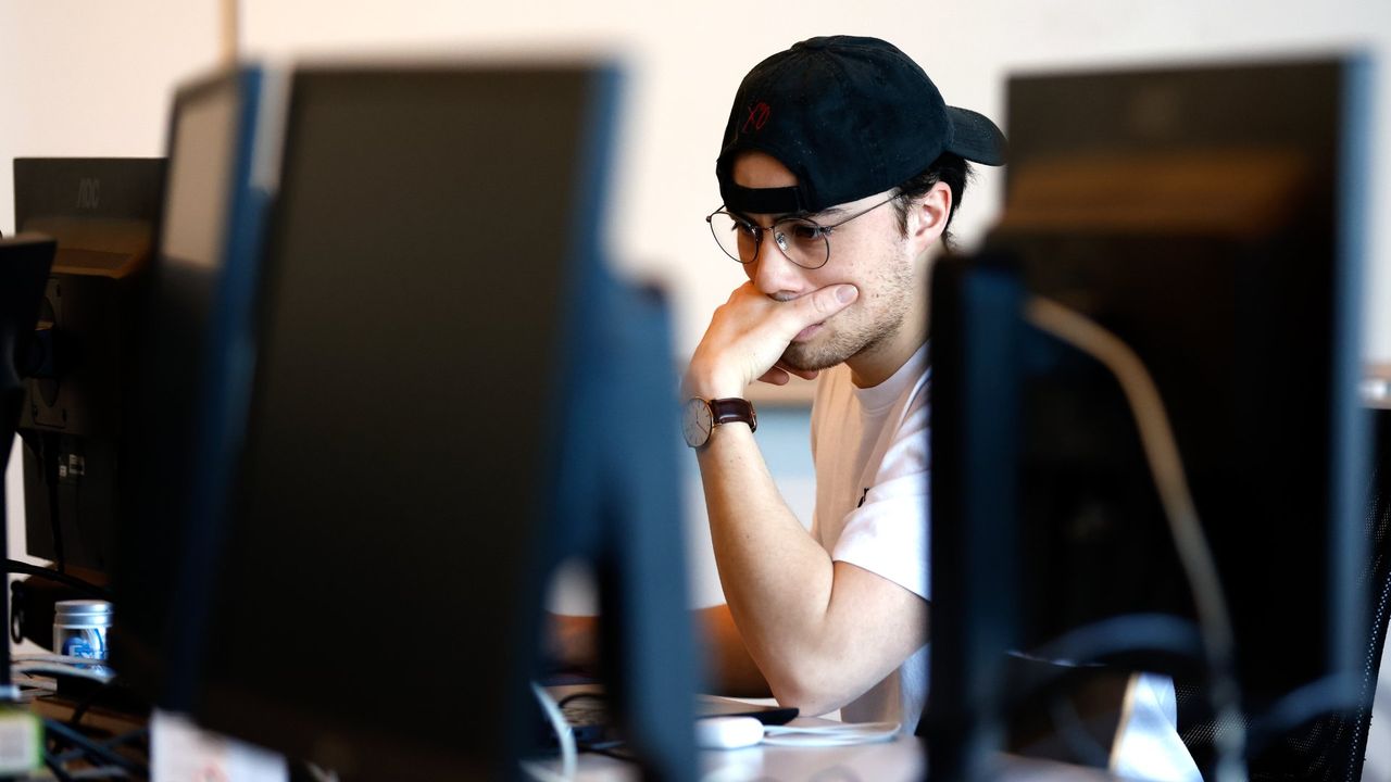 En konsentrert student sitter foran en dataskjerm i et klasserom, omringet av flere skjermer i et fokusert studiemiljø.