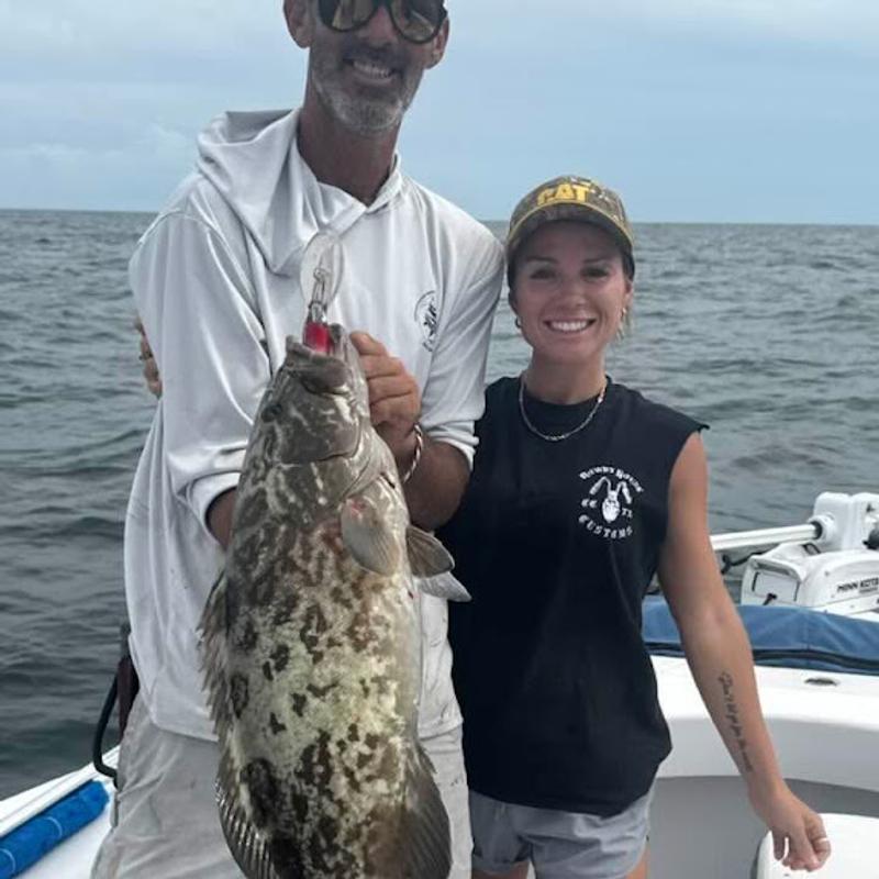 Gag grouper with a diving plug still in its mouth, held up beside a smiling charter guest