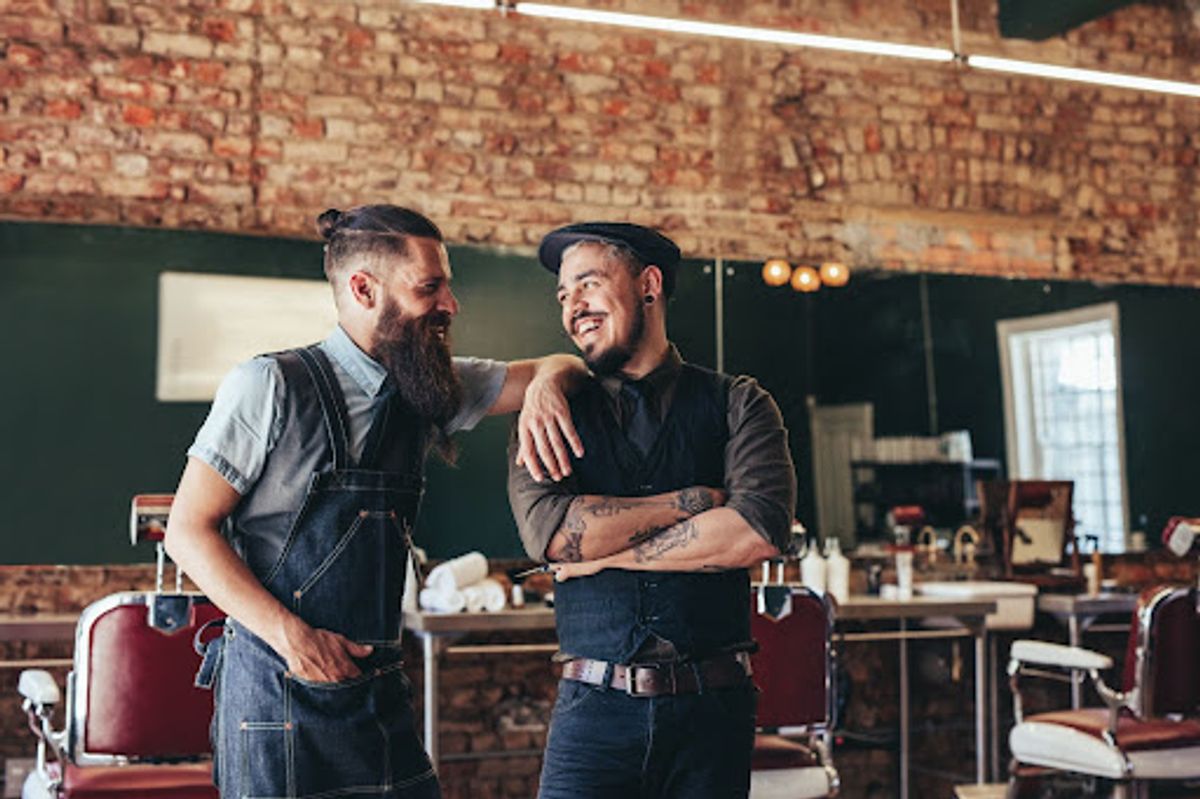 Two barbers smiling and chatting in a rustic barbershop.