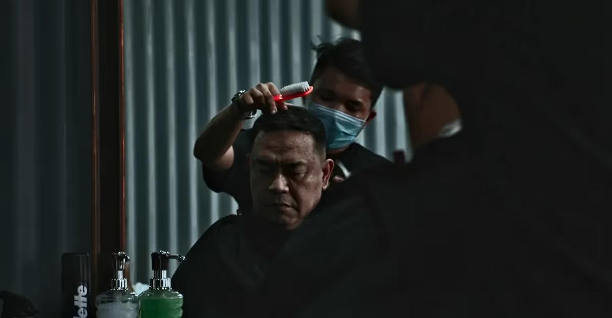 A barber wearing a face mask combs a client's hair during a haircut in a barbershop.