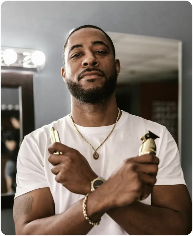 A barber stands in a barbershop holding a trimmer and a straight razor with arms crossed, looking at the camera.