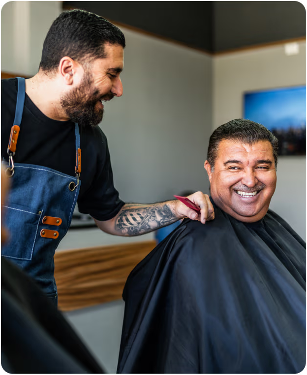 A barber in an apron smiles while trimming a seated client's hair; the client, wearing a black cape, looks over and smiles in a barbershop.