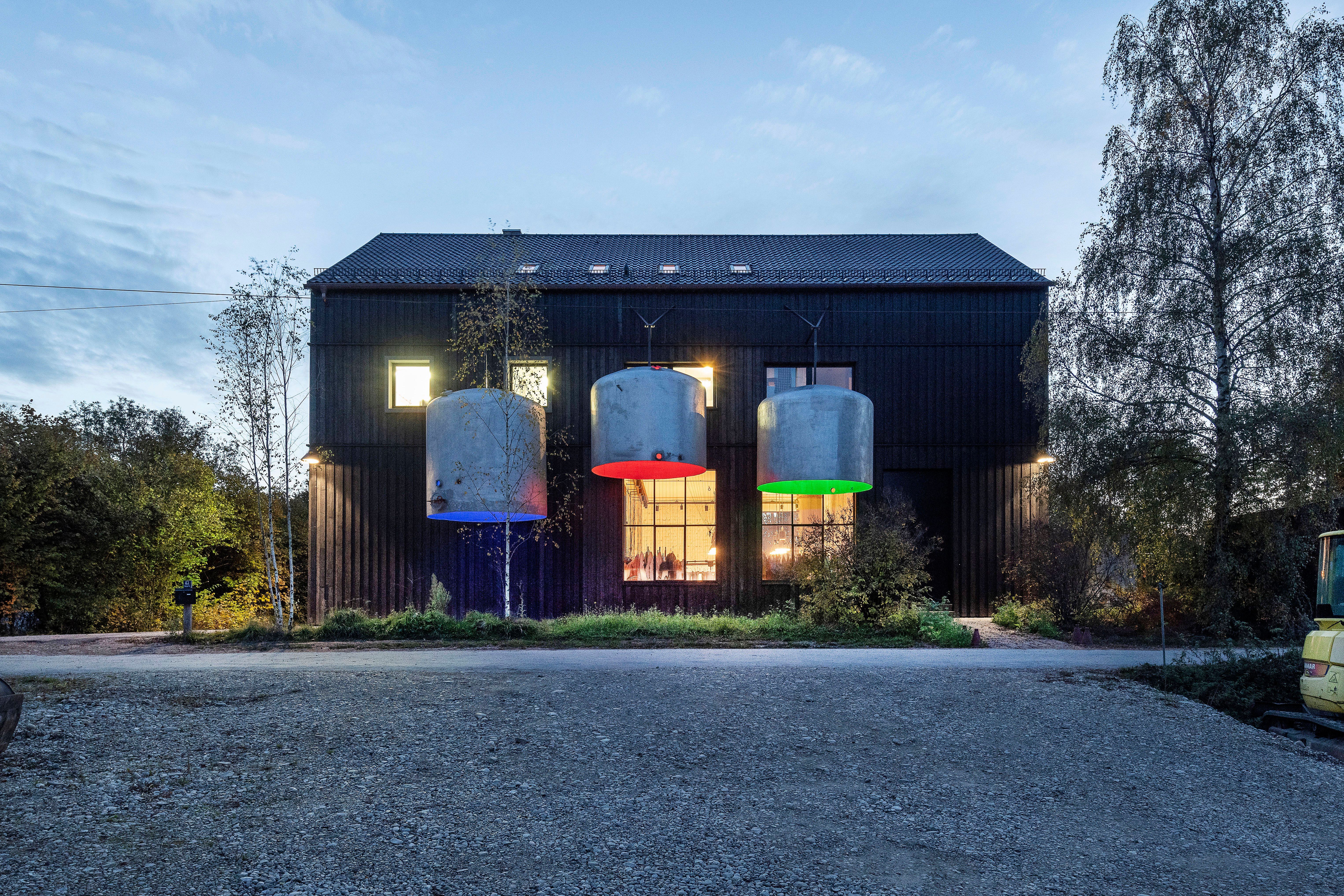 A dark, barn-like house at dusk with three large, cylindrical tanks hanging in front, lit by blue, red, and green lights.