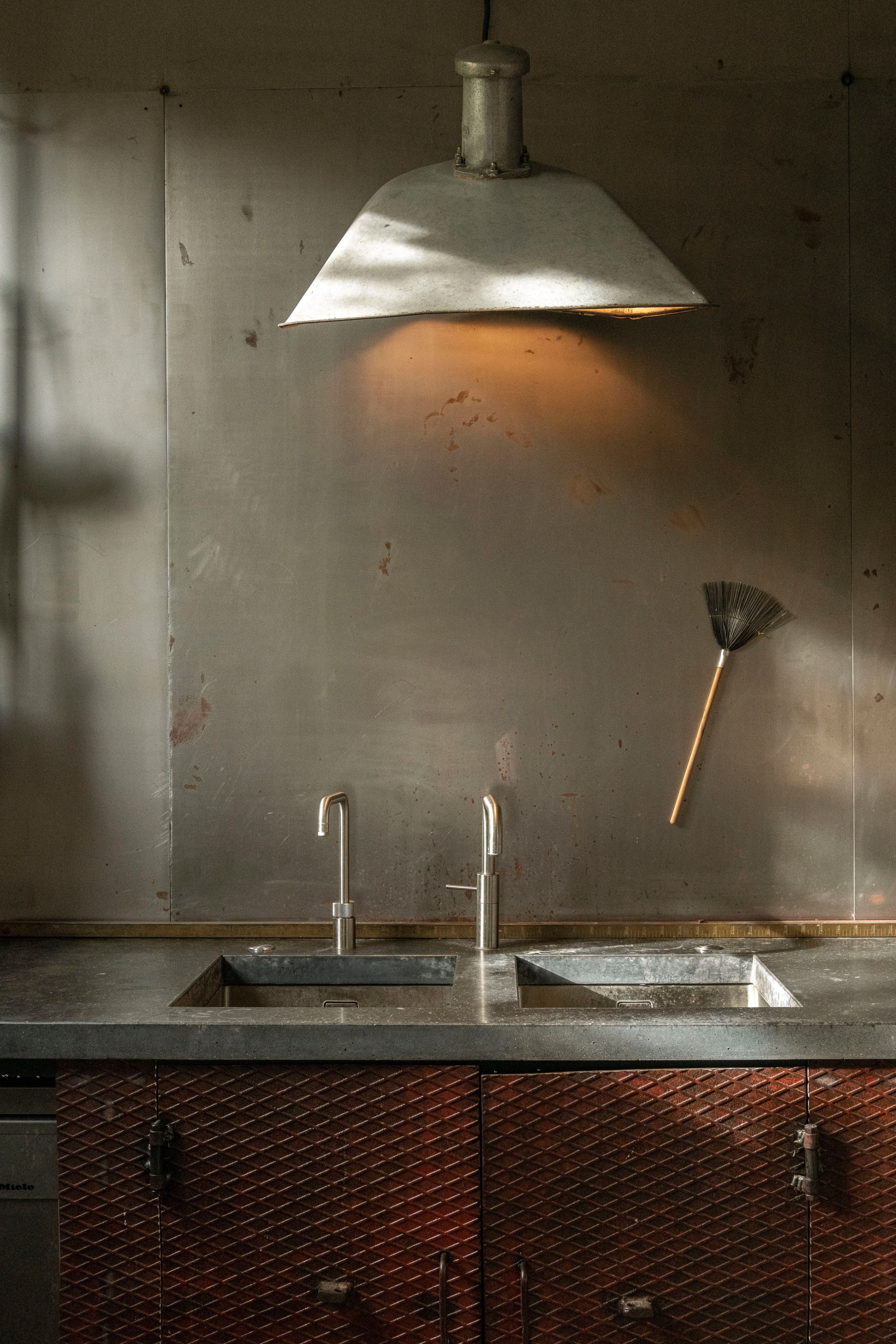 An industrial kitchen with a metal pendant light illuminating a weathered metal backsplash above two sinks and dark red diamond-patterned cabinets.