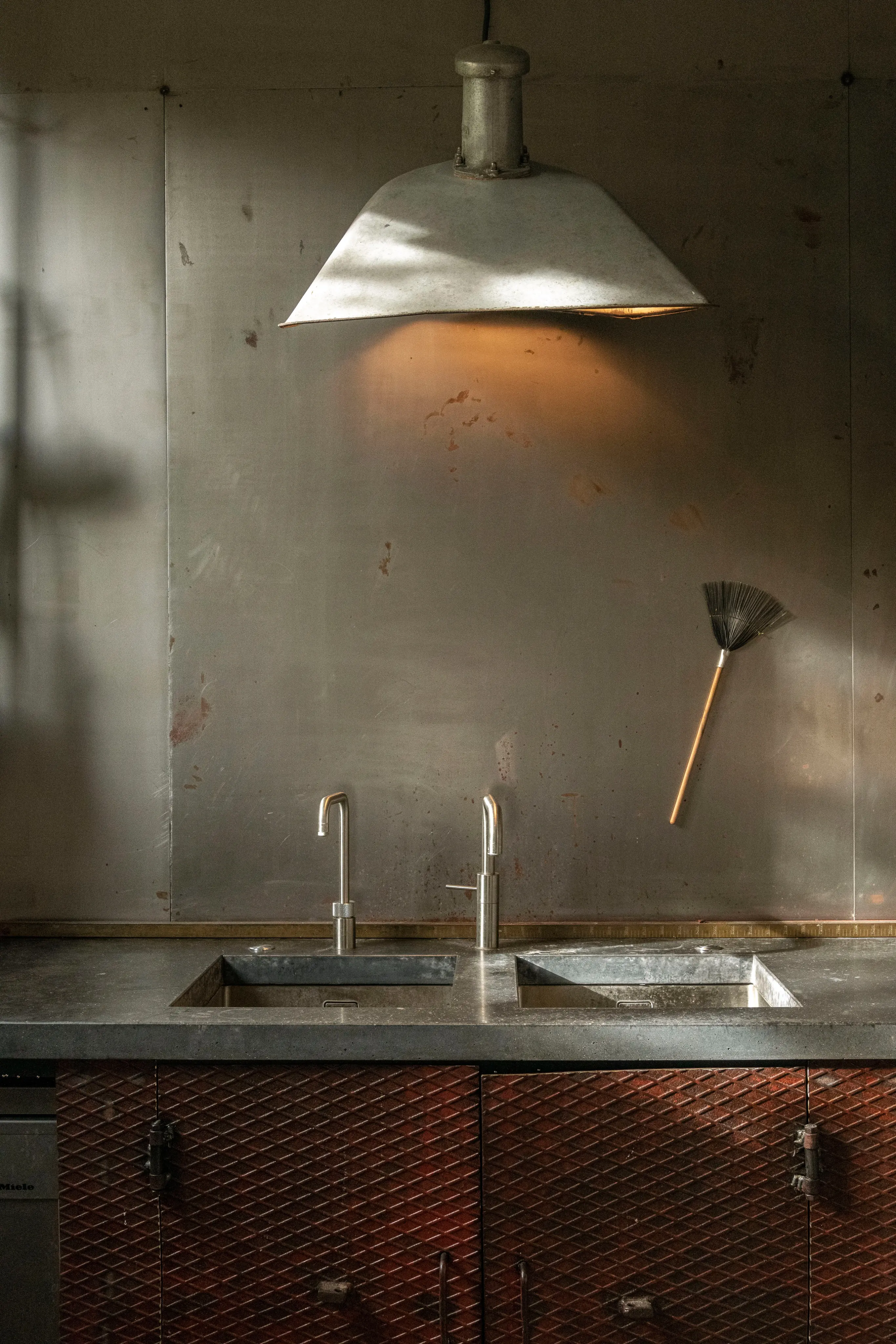 An industrial kitchen with a metal pendant light illuminating a weathered metal backsplash above two sinks and dark red diamond-patterned cabinets.