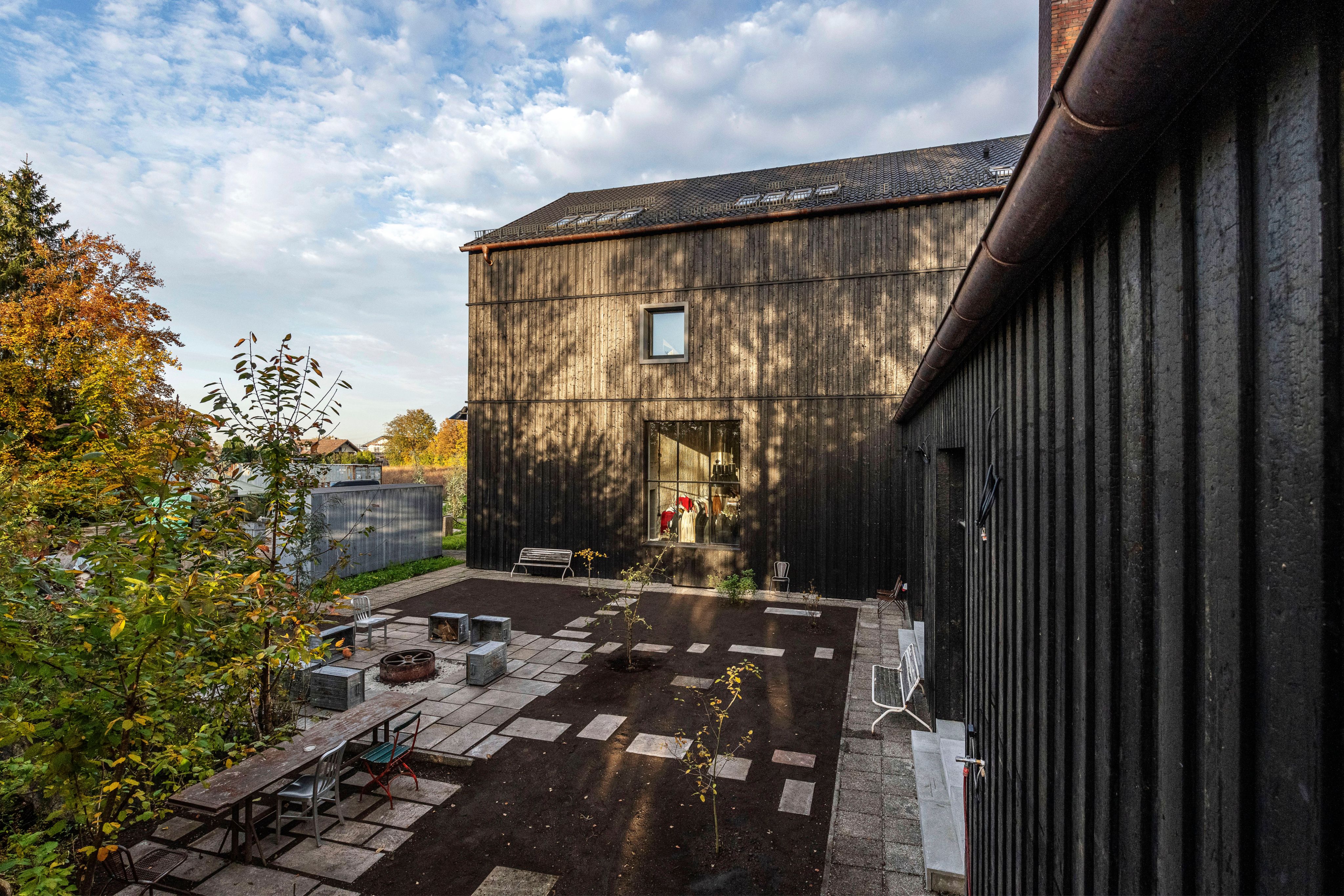 Exterior view of a dark wooden house with a patterned patio, fire pit, and autumn trees.