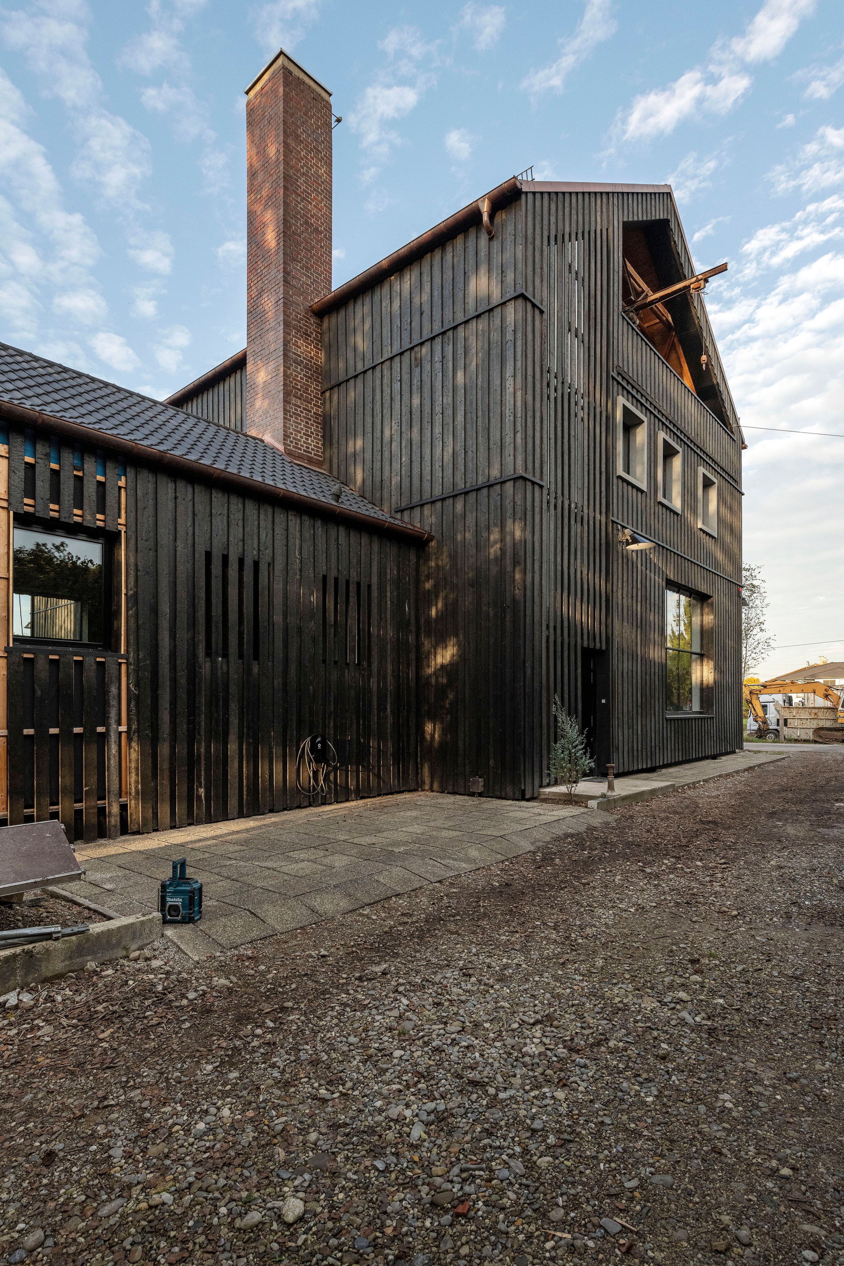 A modern building with dark charred wood siding and a tall brick chimney, beside a gravel path. Its taller section has an open gable.