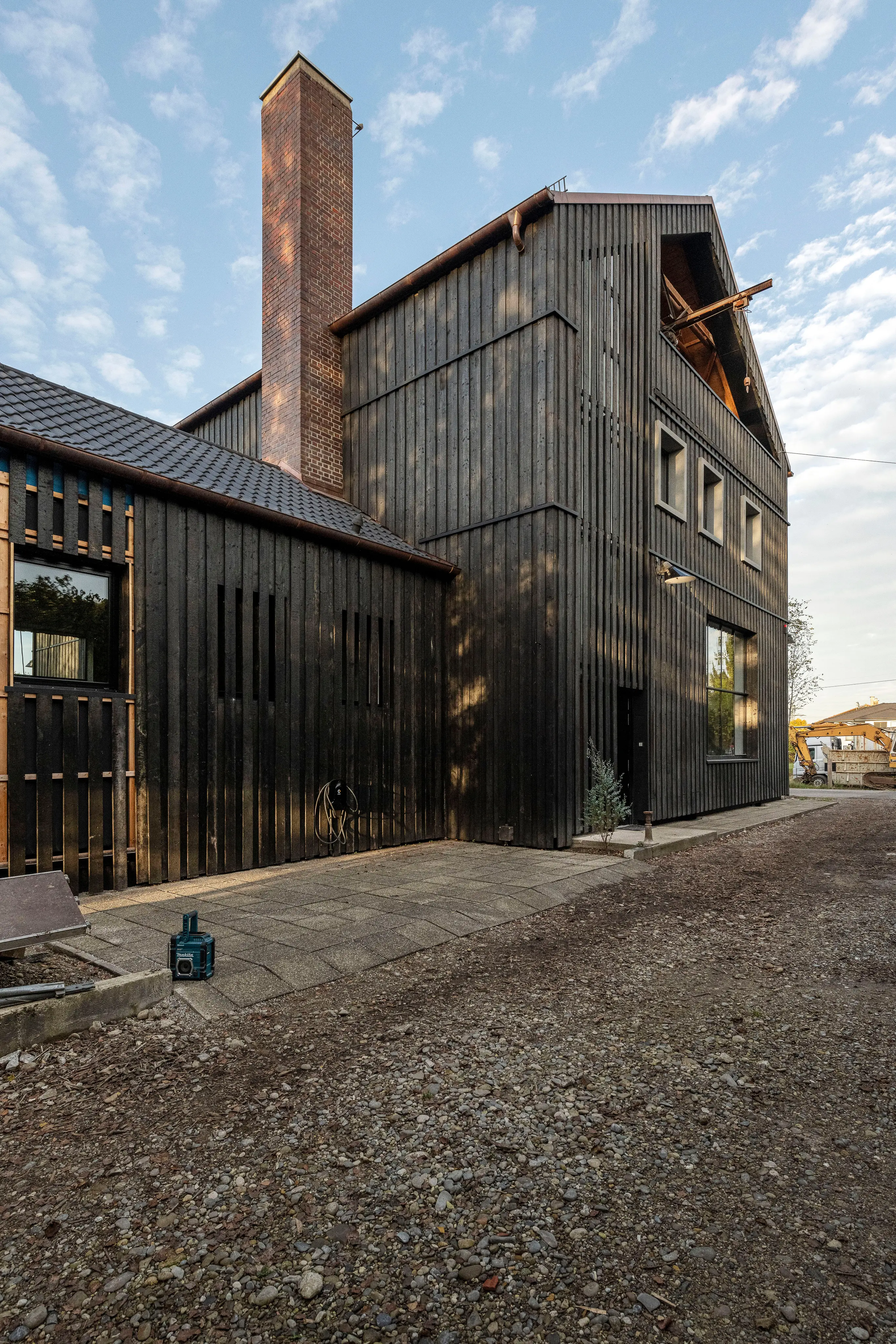 A modern building with dark charred wood siding and a tall brick chimney, beside a gravel path. Its taller section has an open gable.