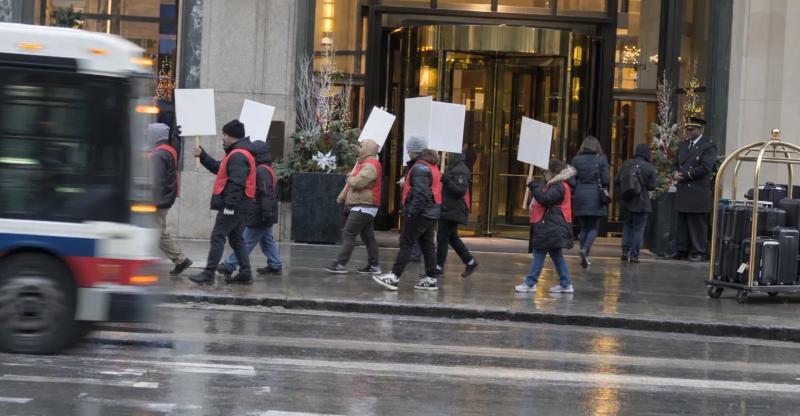 Hotel workers in red vests walk a picket line past a downtown Chicago hotel entrance on a wet winter morning.