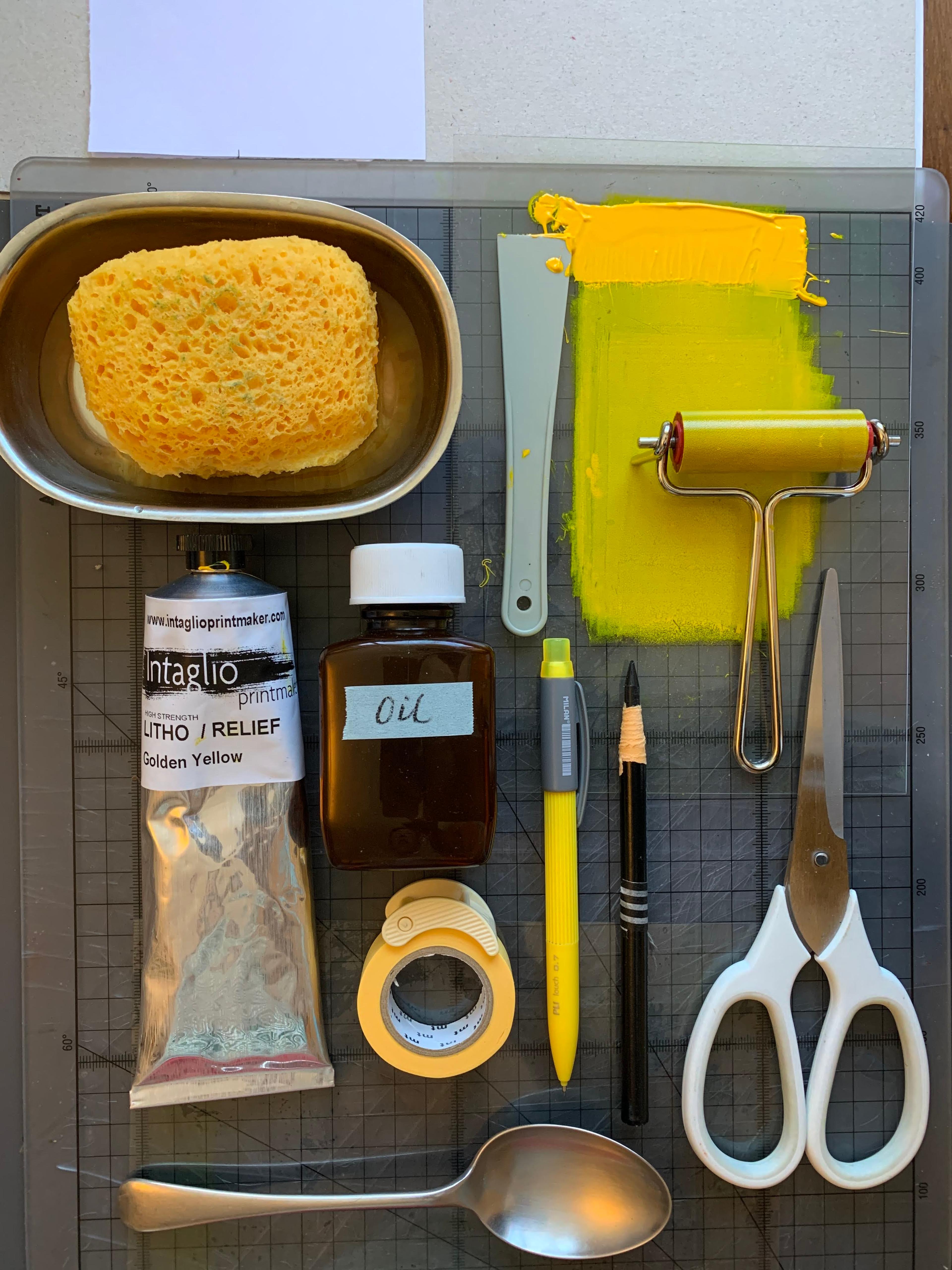 Materials laid out neatly on a table top, including scissors, a spoon, a tube of paint, a bottle of oil, a sponge, a pencil, a pen and a roller with yellow paint on it.