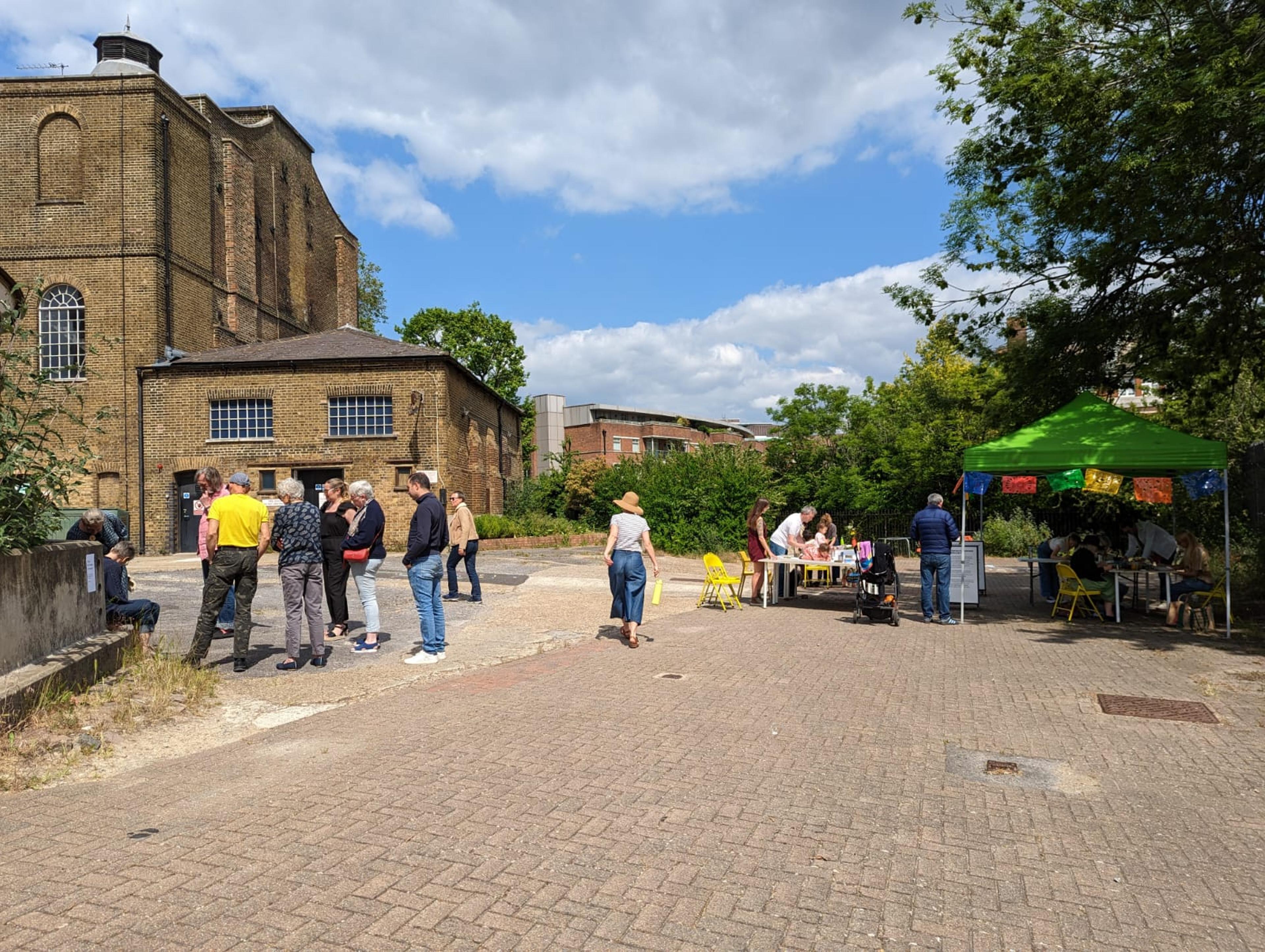 A photo with a building in the background, a gazebo and lots of foliage in the foreground. People stand around.