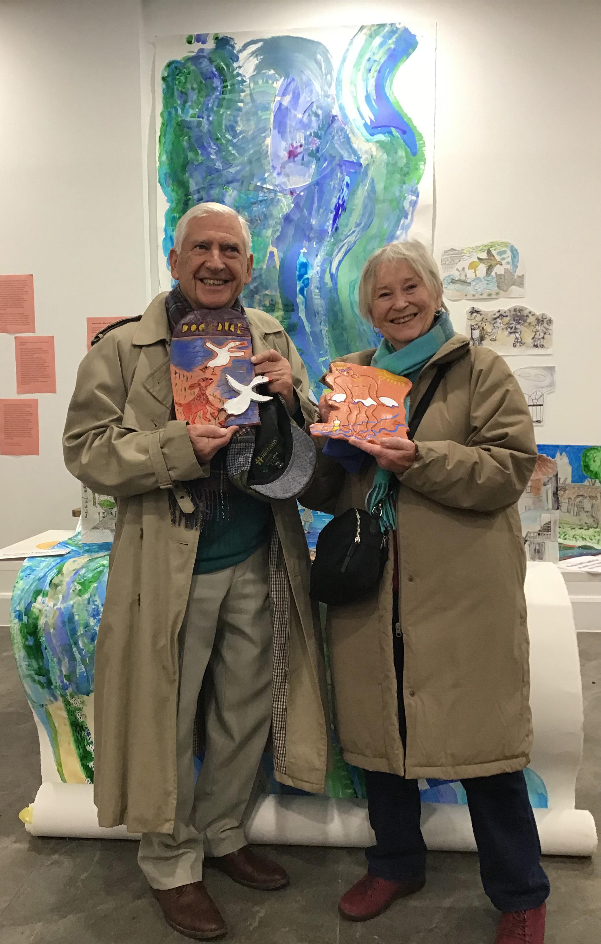 Two people holding ceramics standing in front of a large scroll and illustrations displayed on the wall.