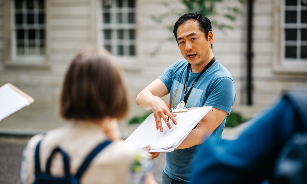 Man in a blue t-shirt points to a drawing on a clipboard while speaking to a person whose back is to the camera.