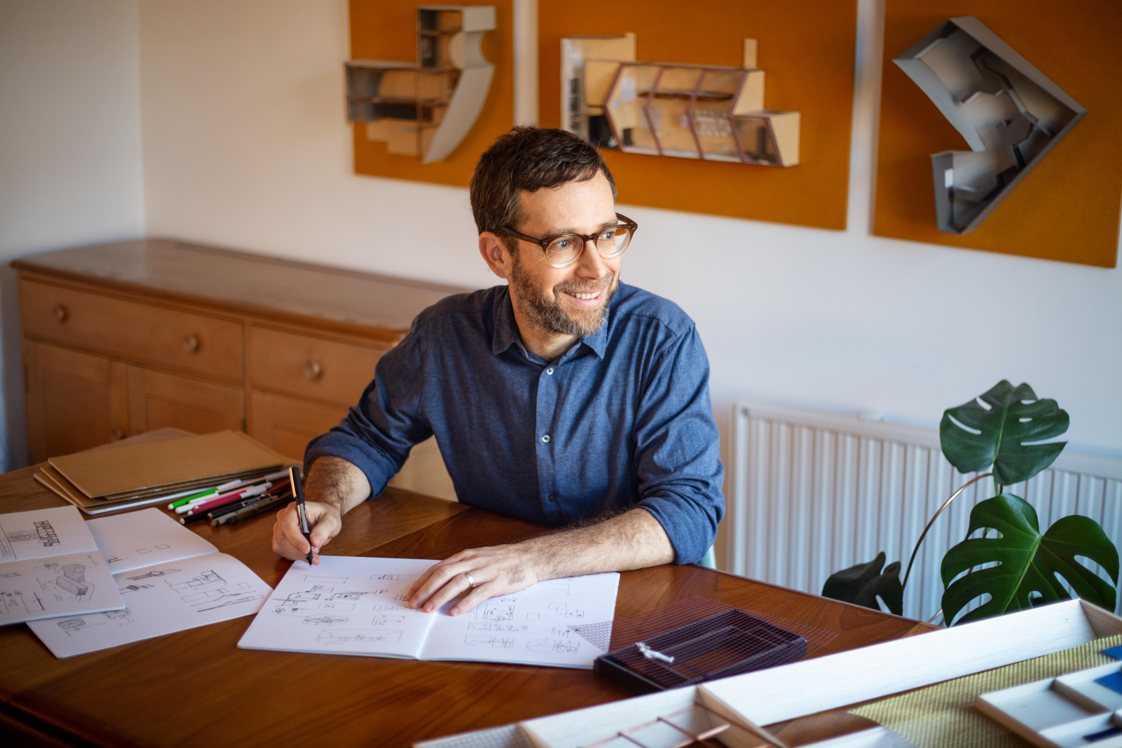 A photograph of a man with glasses sitting at a wooden desk, with open sketchbooks, pens and pencils. In the background some 3D models are displayed on the wall. 