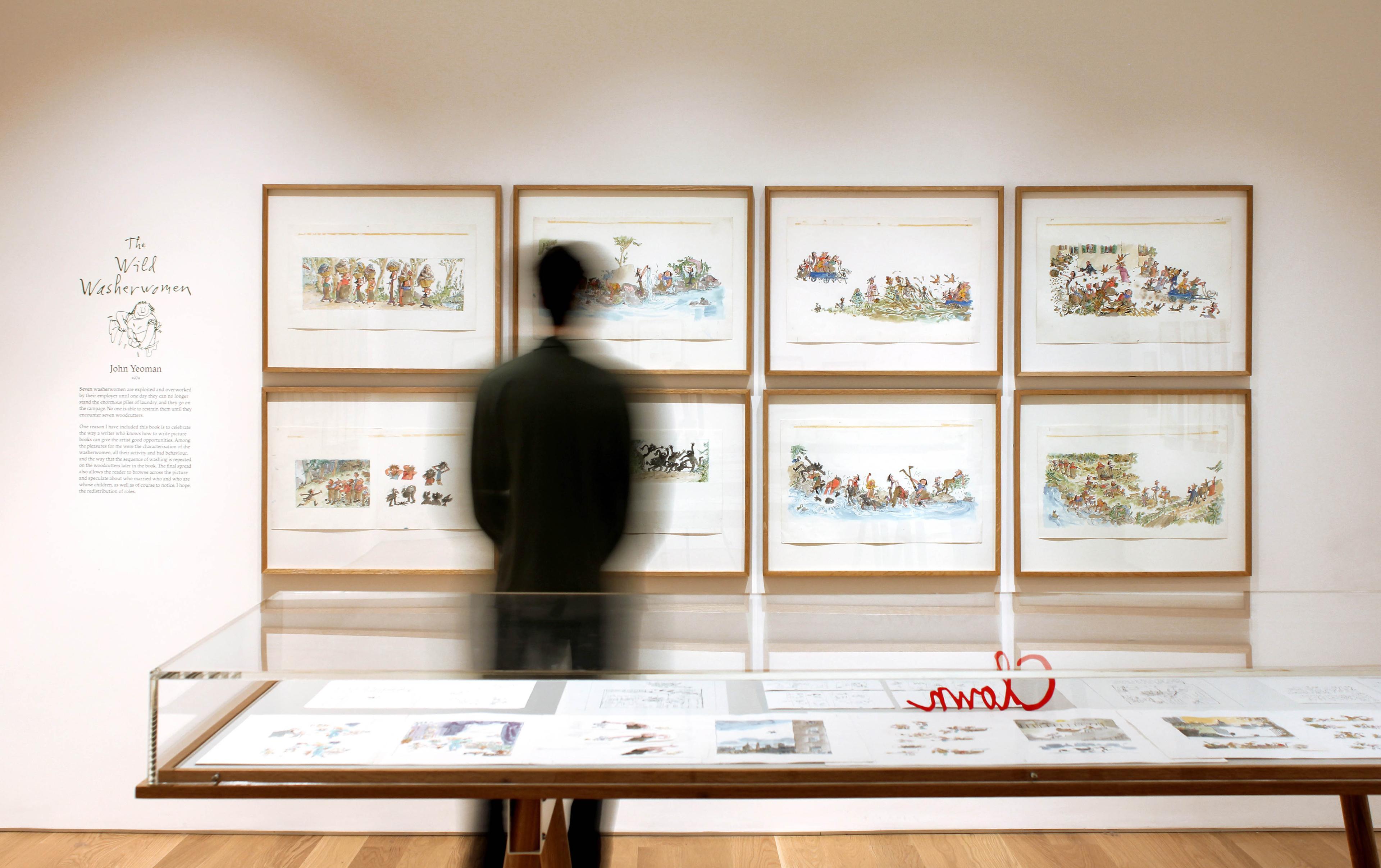 A person in an exhibition space, with framed illustrations on the wall and a glass display cabinet case.