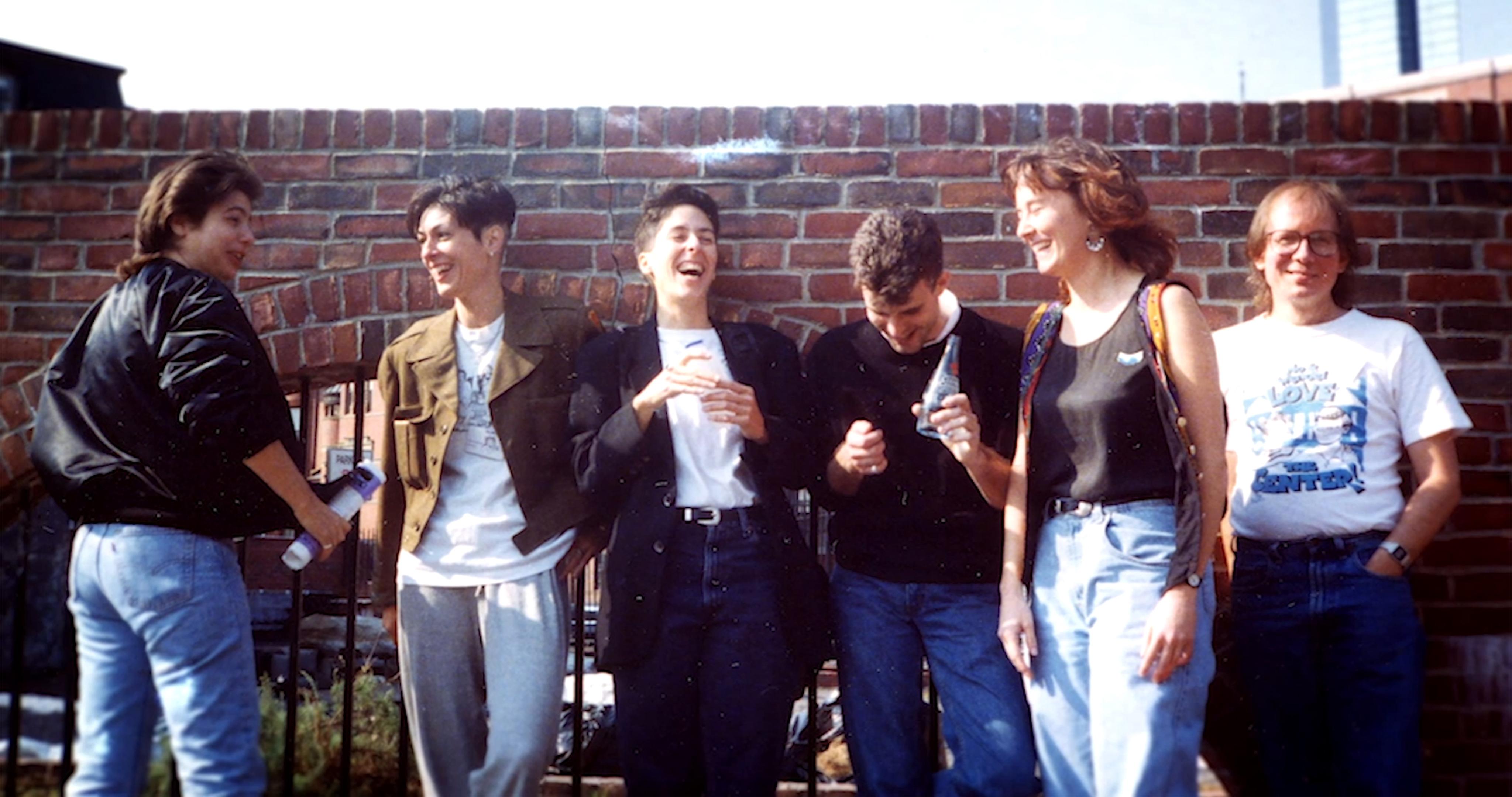 A group of people stand relaxed and laughing against a brick wall.
