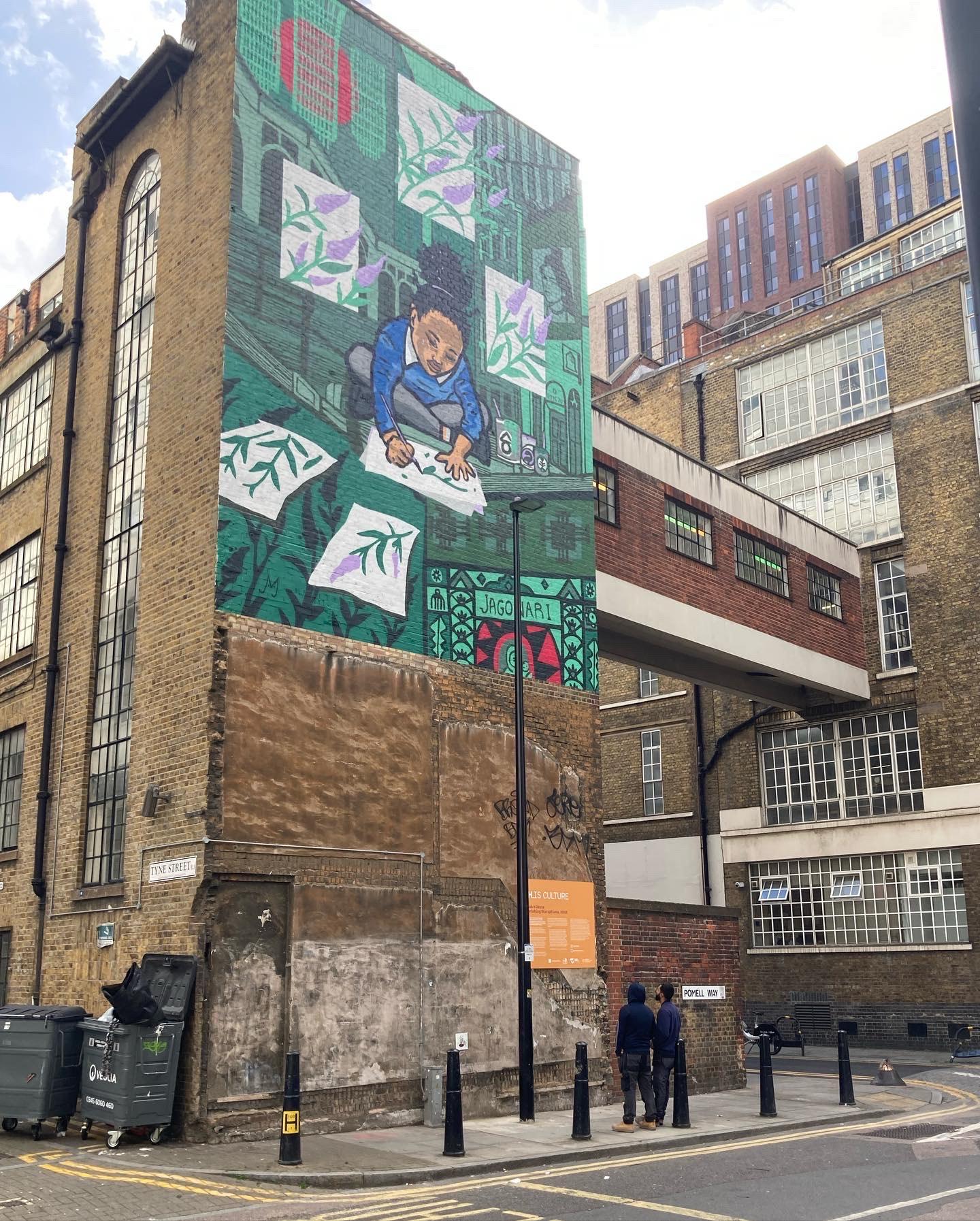Photograph of the side of a tall brick building, surrounded by other buildings, with a painted mural of a small child sitting cross-legged drawing flowers on sheets of paper, with some of them featured around here. The background is a green scene.