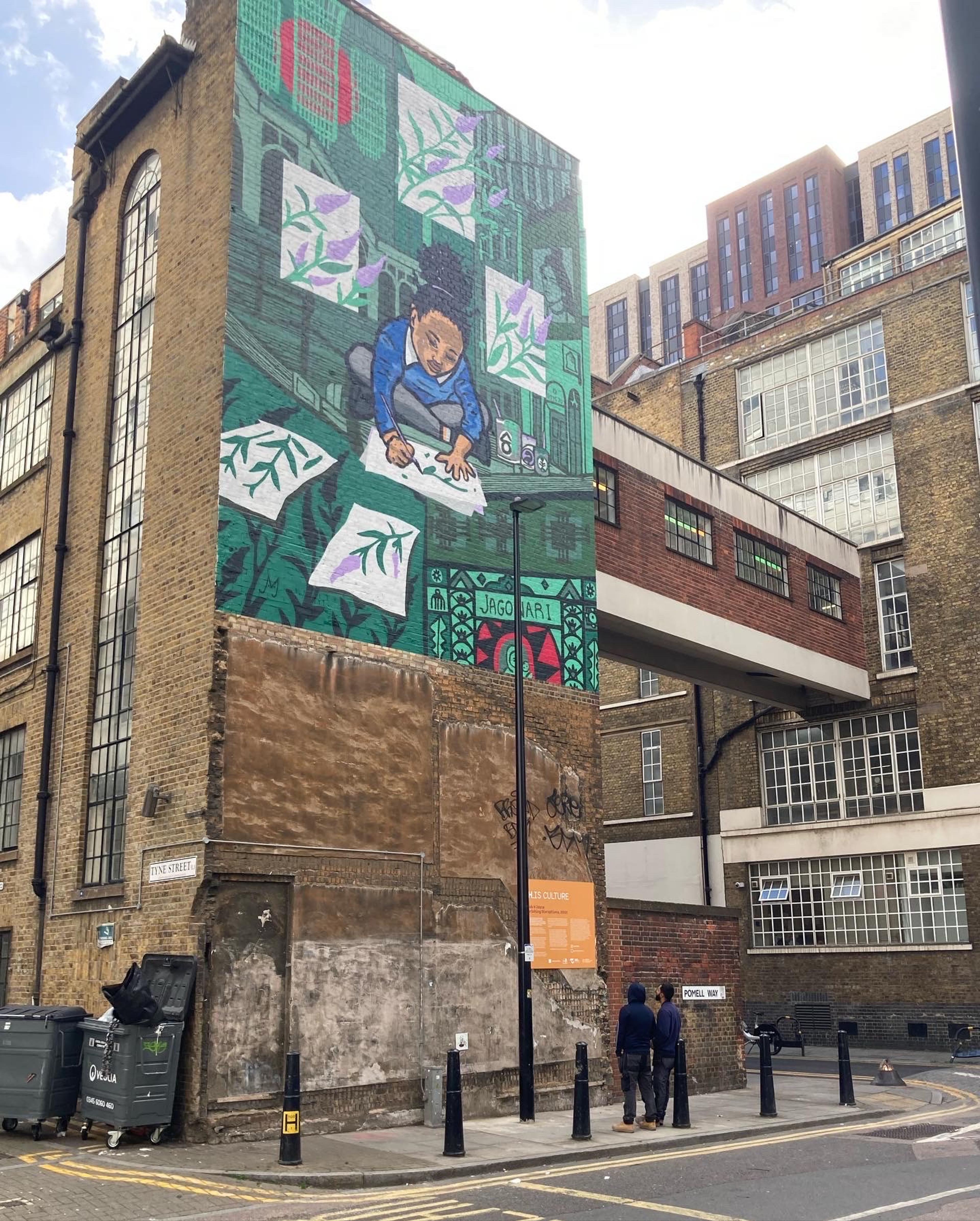 Photograph of the side of a tall brick building, surrounded by other buildings, with a painted mural of a small child sitting cross-legged drawing flowers on sheets of paper, with some of them featured around here. The background is a green scene.