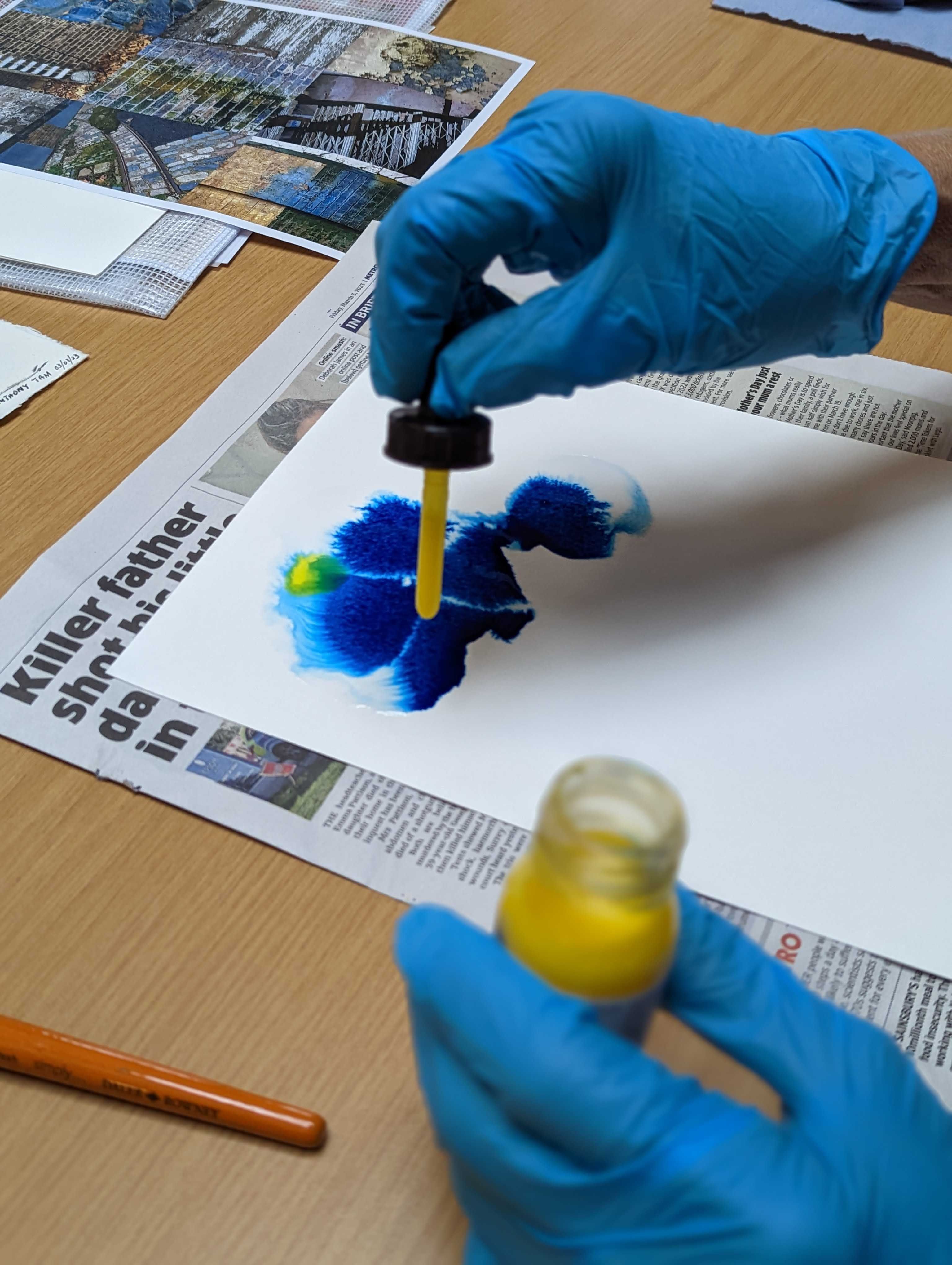 Two gloved hands, one hand is using an ink dropper to drop ink onto some paper.