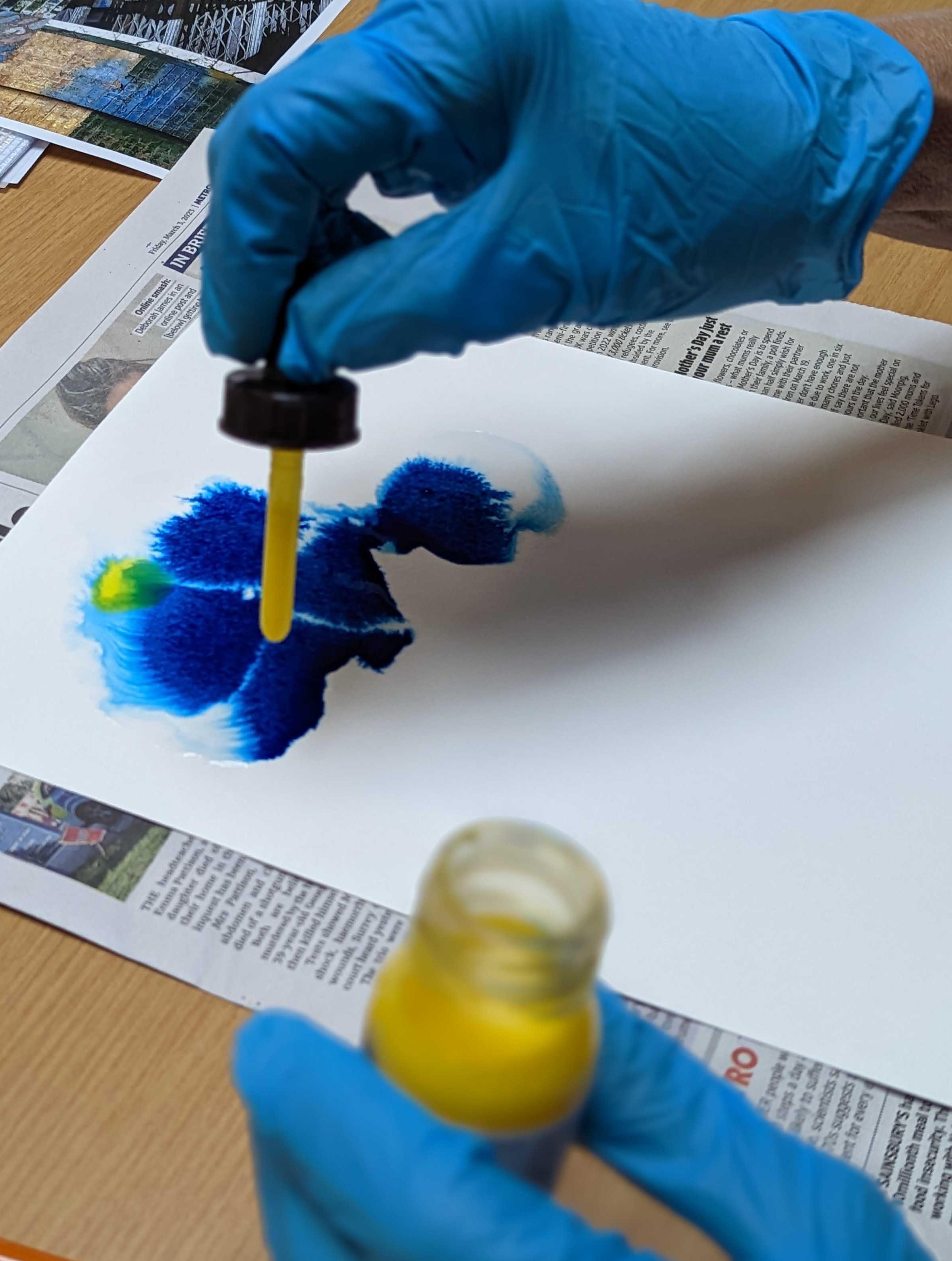 Two gloved hands, one hand is using an ink dropper to drop ink onto some paper.
