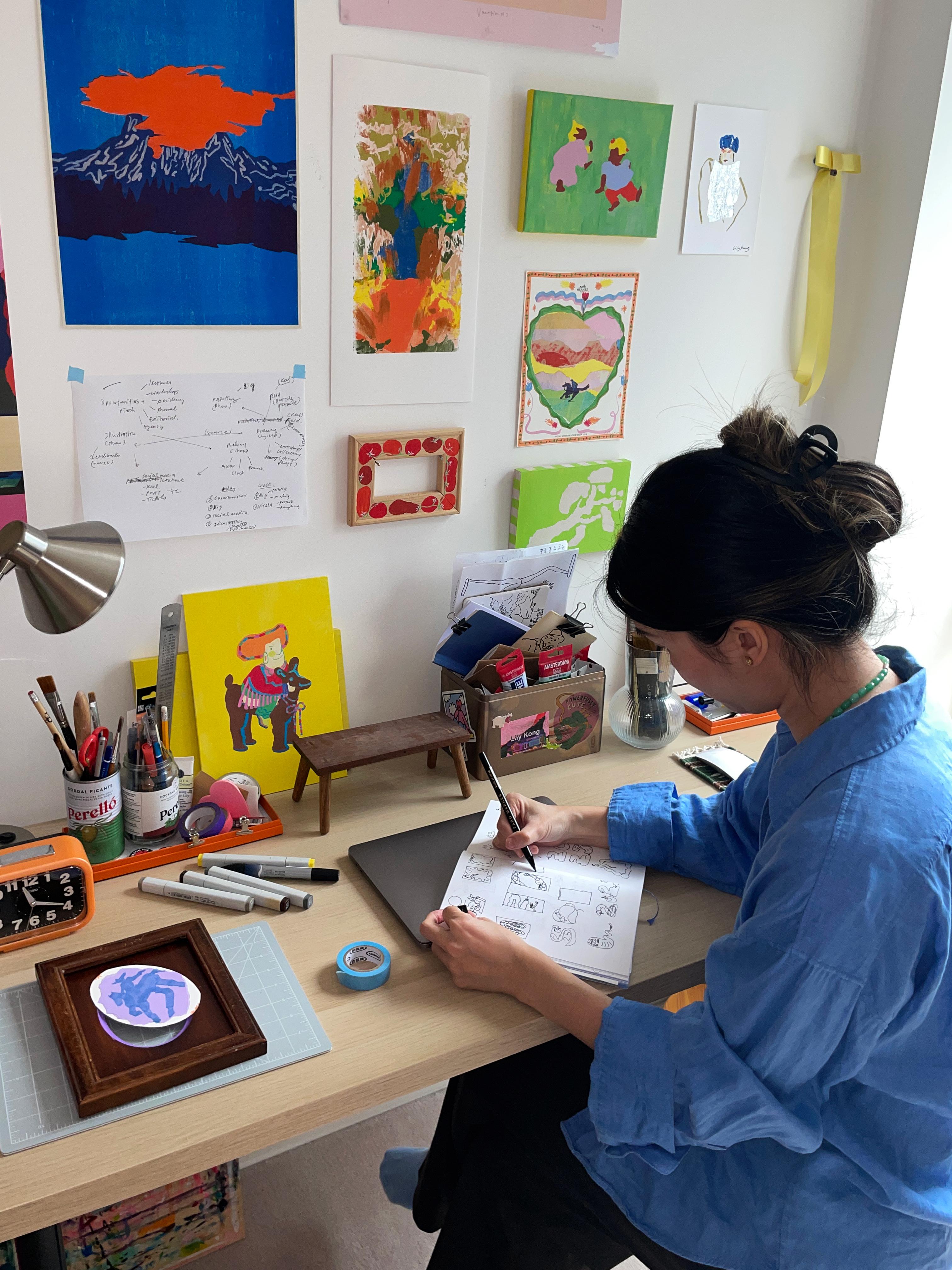 A woman in a blue shirt sits at a desk drawing. The walls have illustrated materials on them and there are creative materials on the desk.